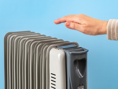 The top of a silver space heater with a hand over it feeling the heat against a light blue wall